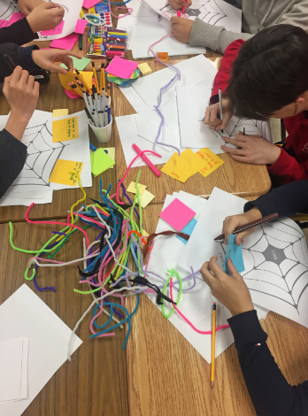 Several people sit around tables talking with each other, interacting on digital devices and writing on pads of paper and sticky notes.